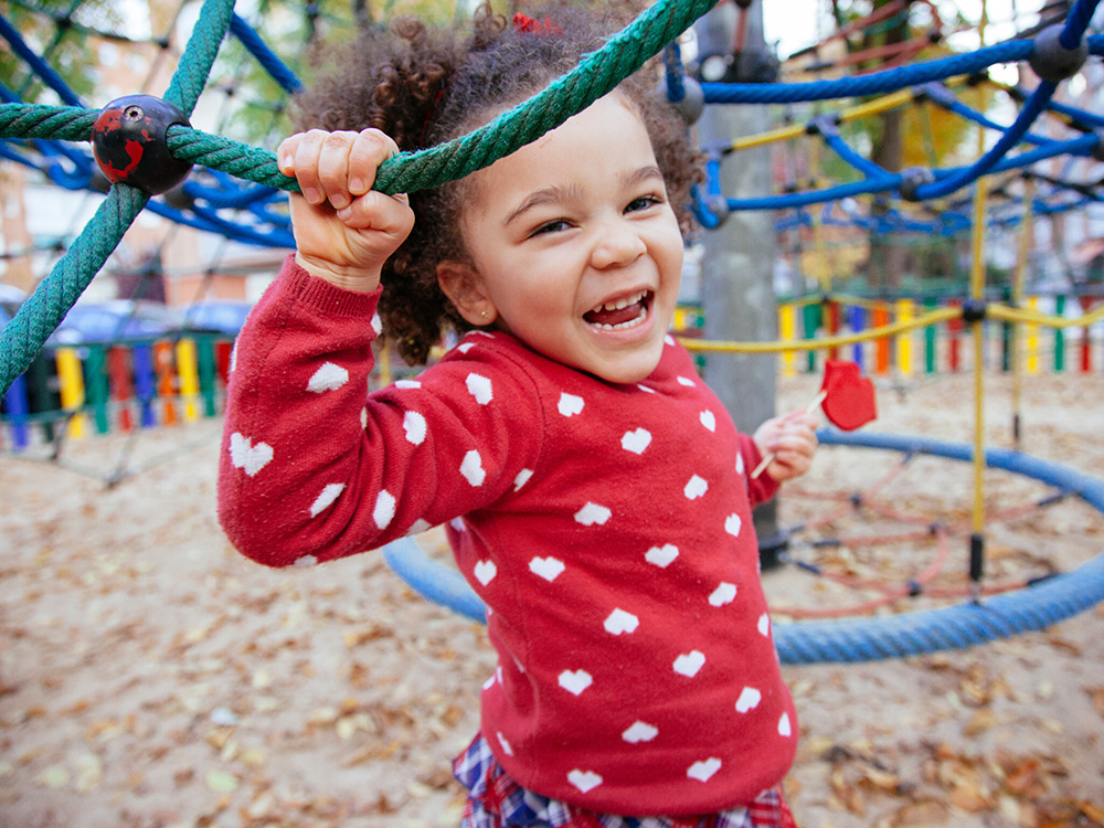 A cheerful girl on a playground, smiling widely while holding onto a climbing structure.