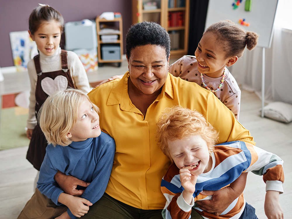 A smiling teacher surrounded by four kids, all joyfully interacting and having fun together.