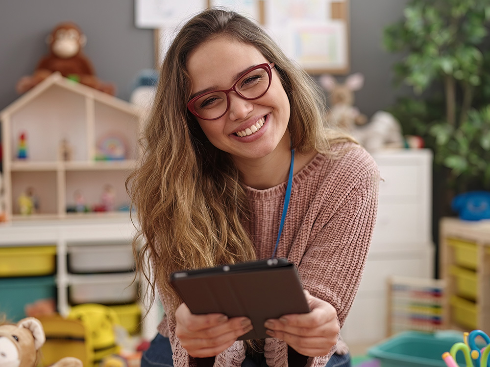 A young teacher with glasses, smiling and holding a tablet, in a playful environment.