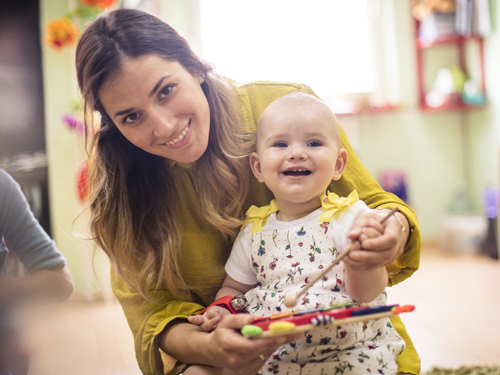 A smiling baby, with the teacher holding a colorful instrument.
