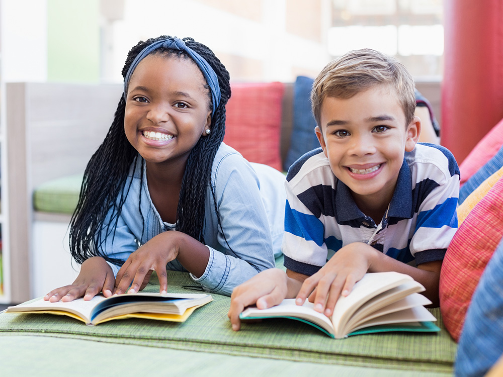 Two children lying on a couch, both happily enjoy the reading of books.