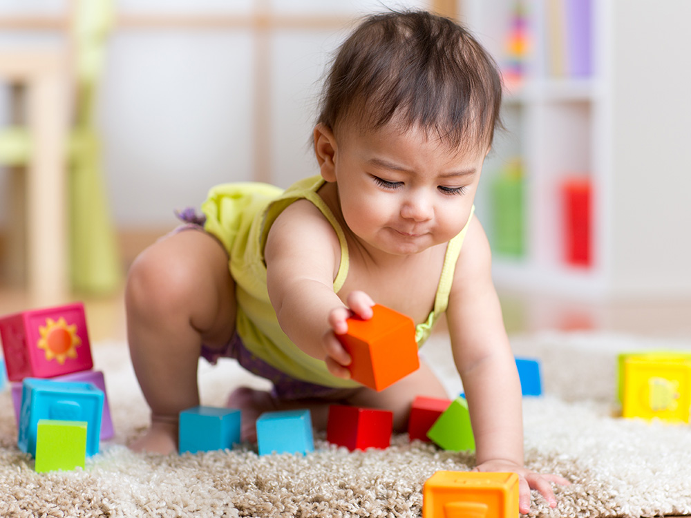A baby reaching for colorful building blocks while sitting on a carpet.