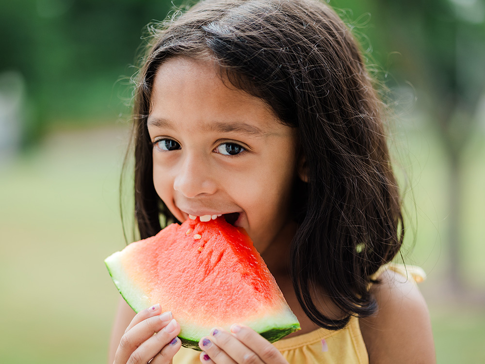 A girl with short hair, happily eating a slice of watermelon, enjoying a sunny day outside.