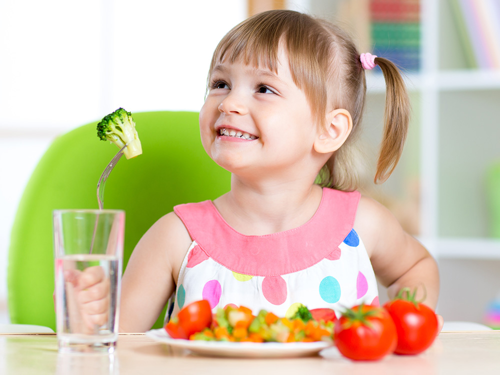 A girl happily holding a piece of broccoli with a glass of water nearby, smiling at her meal.