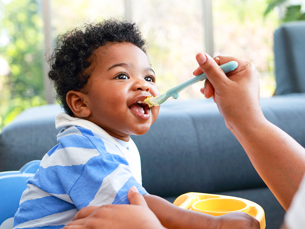 A baby being fed by a teacher, looking up happily at the person feeding him with a spoon.