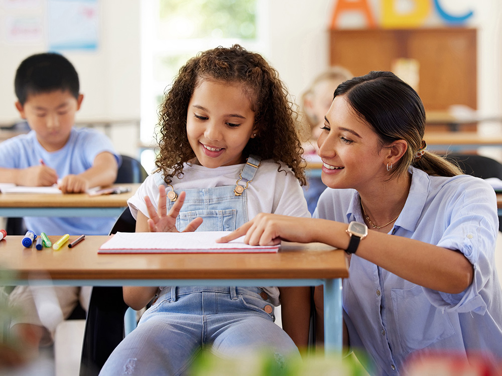 A girl and a teacher smiling while reviewing a notebook, with other students working in the background.