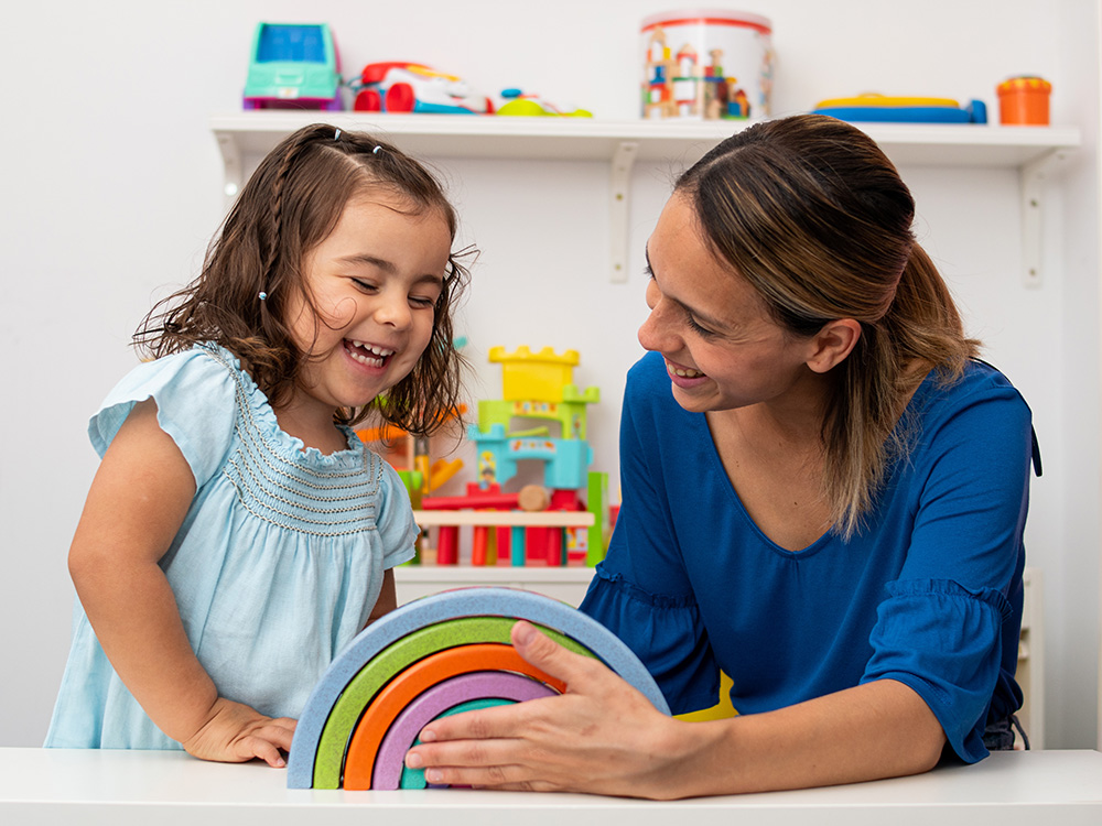 A teacher and a student girl smiling, playing with colorful stacking rings at the table.