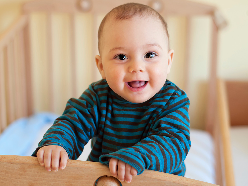 A smiling baby in a striped shirt, holding onto the edge of a crib.
