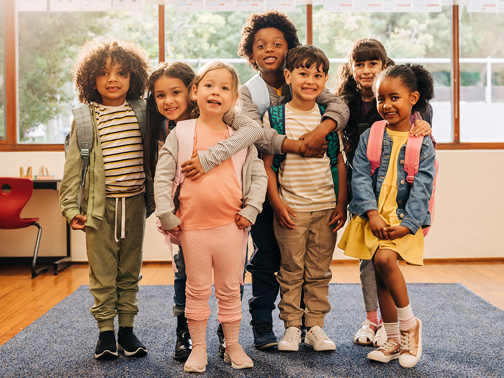 A group of seven smiling children posing together, all wearing backpacks in a bright classroom setting.