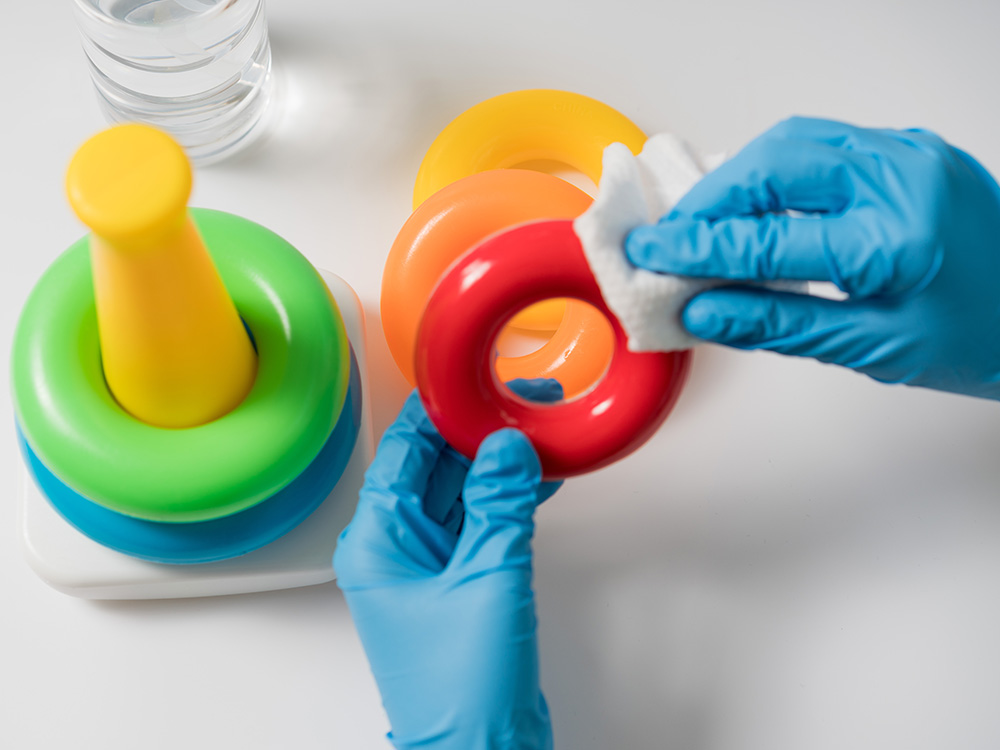 A pair of hands wearing blue gloves cleaning a red toy ring from a colorful stacking toy set.