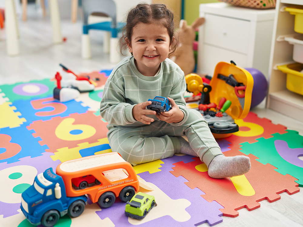 A smiling toddler sitting on a colorful play mat, holding a toy car while surrounded by various toys.