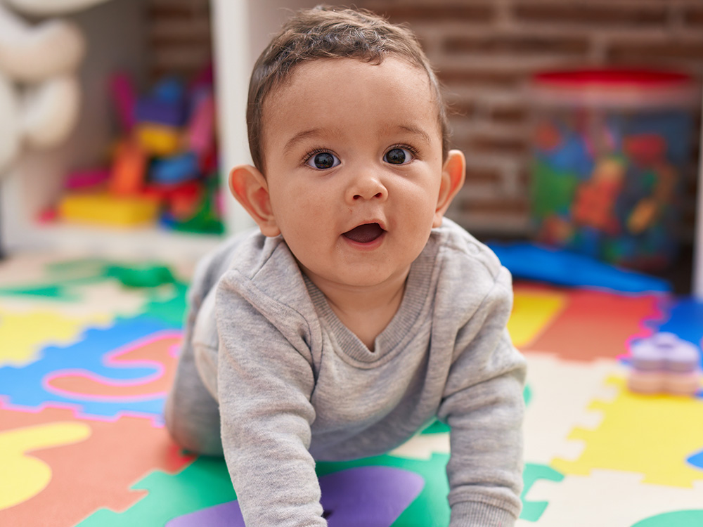 A baby crawling, looking surprised and happy, wearing a gray outfit.