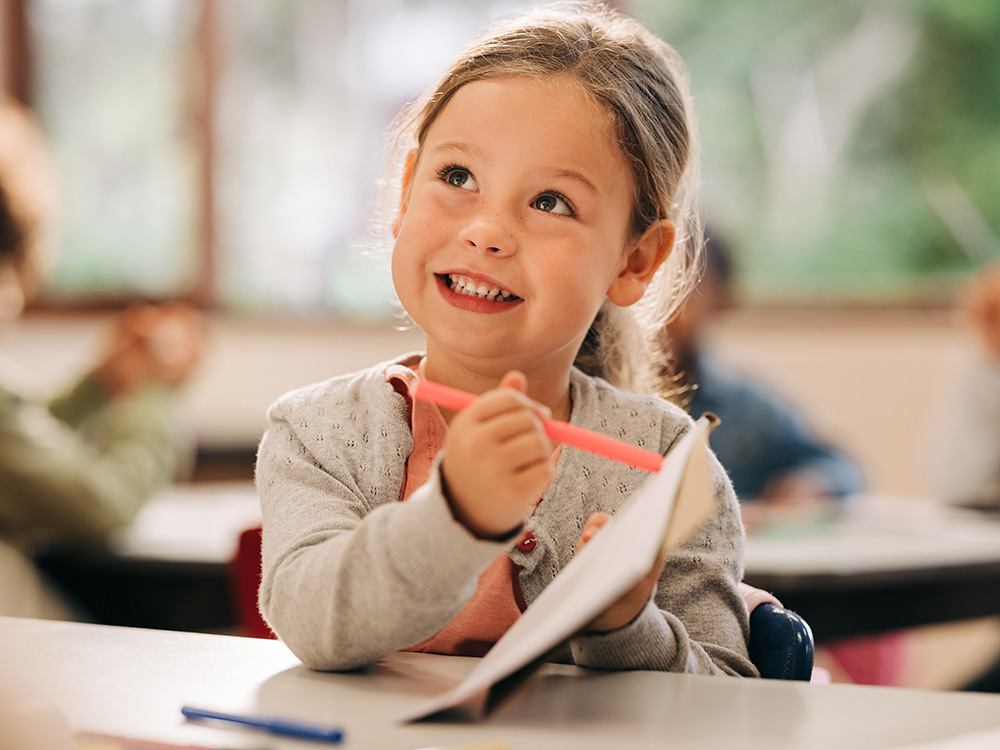 A smiling girl in a classroom, looking up with a pink pen in her hand, engaged in her work.