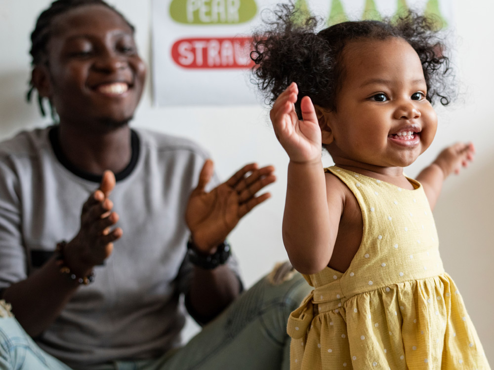 A smiling toddler in a yellow dress, dancing, with a teacher clapping and encouraging her in the background.