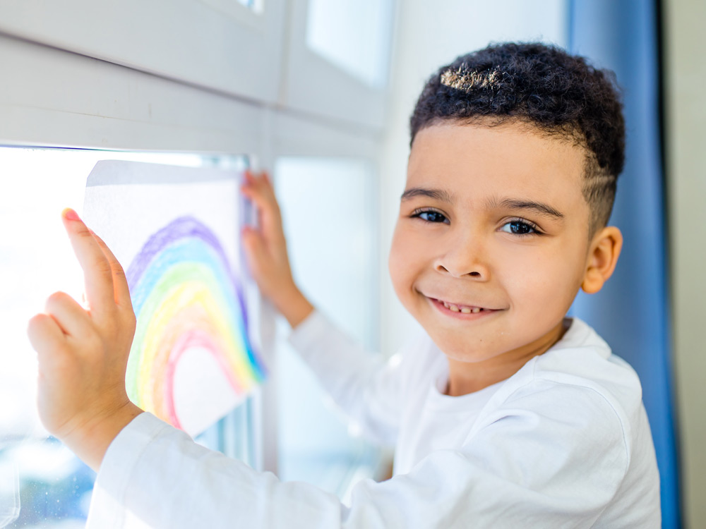 boy smiling while putting a colorful drawing of a rainbow on a window.