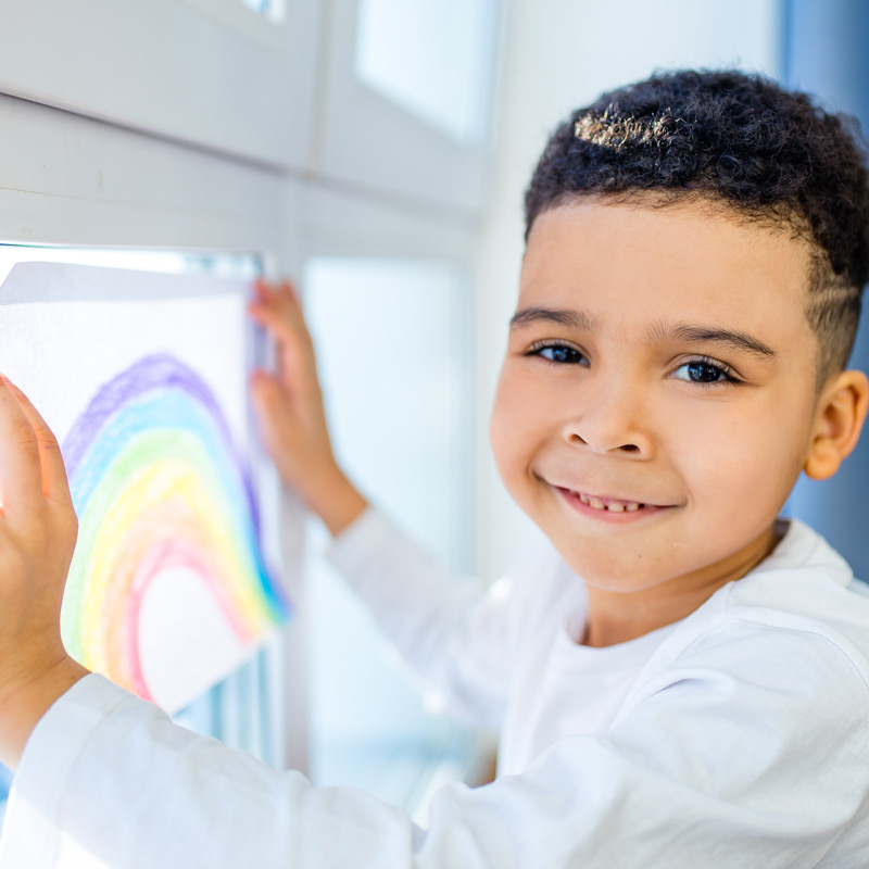 boy smiling while putting a colorful drawing of a rainbow on a window.
