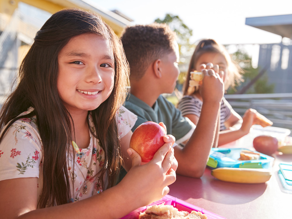 A girl holding an apple while enjoying lunch with friends at a table, all looking engaged and happy.