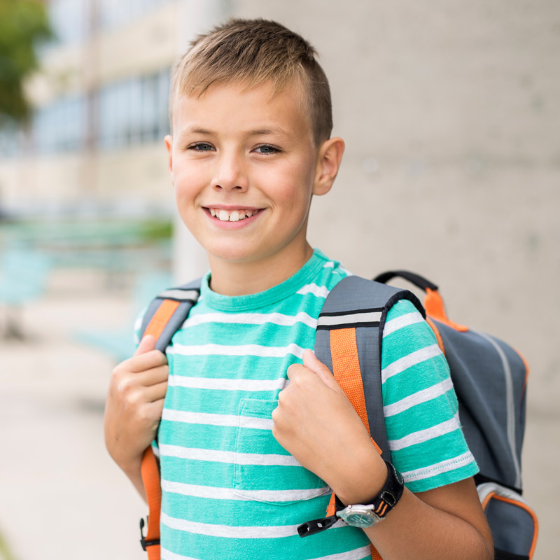 A smiling boy with short hair smile widely, wearing a striped shirt and backpack, standing outside the school.