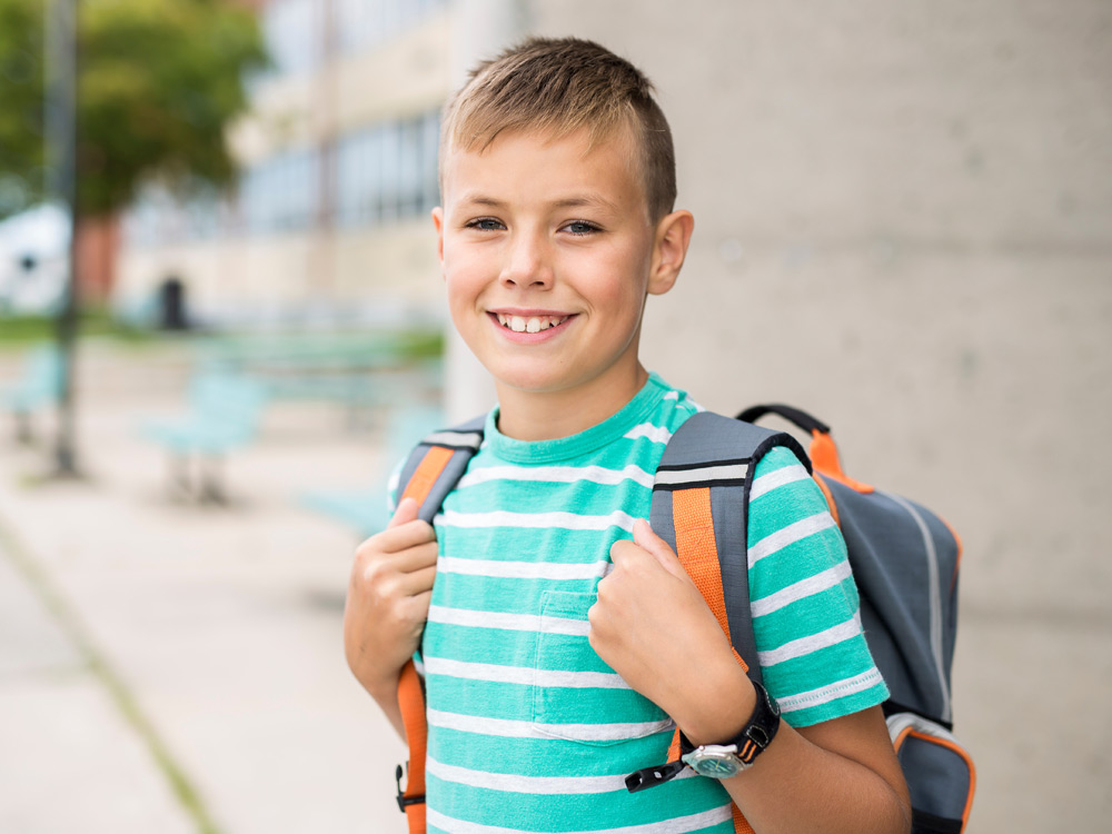 A smiling boy with short hair smile widely, wearing a striped shirt and backpack, standing outside the school.